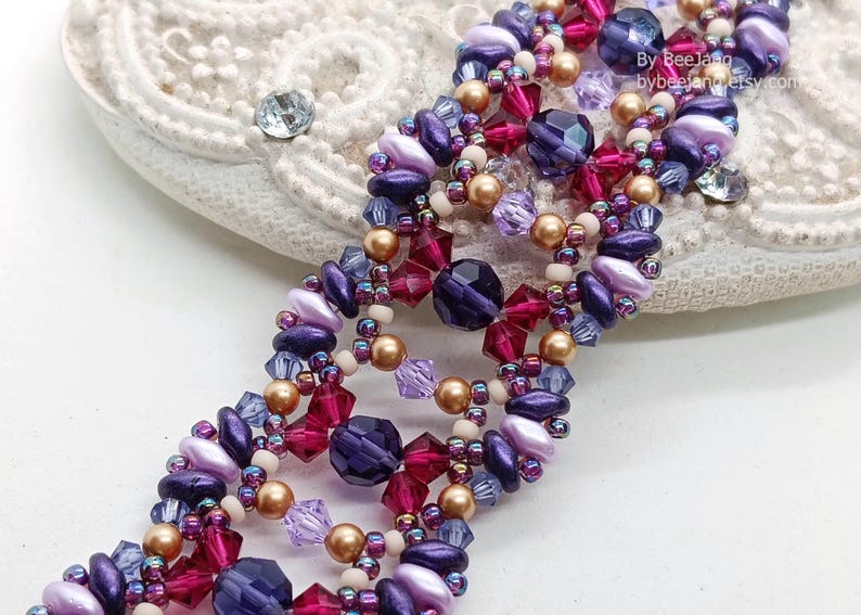 Puede incluir: Una pulsera de cuentas con un dise&ntilde;o complejo, con cuentas moradas, rosas y doradas. La pulsera tiene una construcci&oacute;n de m&uacute;ltiples hilos con varias formas y tama&ntilde;os de cuentas. Las cuentas est&aacute;n dispuestas en un patr&oacute;n repetitivo, creando un accesorio texturizado.