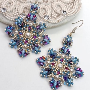 May include: A pair of snowflake-shaped earrings. Each earring features a central pearl surrounded by a pattern of blue, purple, and pink beads. The earrings have silver-colored hooks and are set against a white background.