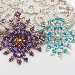 Two handcrafted, intricate beaded snowflake pendants or ornaments displayed on an ornate white surface. The left snowflake features purple, lavender, and gold beads with a central gold pearl, while the right snowflake features white, turquoise, and mint green beads with a central white pearl.