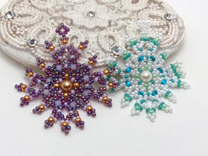 Two handcrafted, intricate beaded snowflake pendants or ornaments displayed on an ornate white surface. The left snowflake features purple, lavender, and gold beads with a central gold pearl, while the right snowflake features white, turquoise, and mint green beads with a central white pearl.