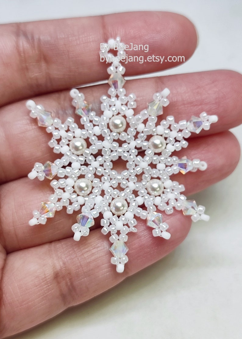 A single, handcrafted, intricate beaded snowflake pendant or ornament featuring white seed beads, clear iridescent crystals, and cream faux pearls, being held in a person's hand.