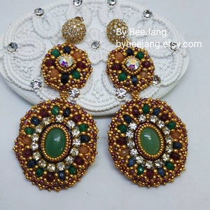 A pair of large, intricate, handcrafted drop earrings featuring an oval green aventurine center stone and extensive bead embroidery using gold, red, dark blue, green, white, and peach-colored seed beads and crystals, displayed on a white decorative surface.
