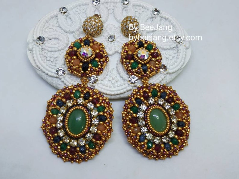 A pair of large, intricate, handcrafted drop earrings featuring an oval green aventurine center stone and extensive bead embroidery using gold, red, dark blue, green, white, and peach-colored seed beads and crystals, displayed on a white decorative surface.
