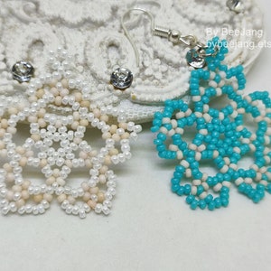 A pair of intricate, handcrafted jewelry pieces, likely earrings, featuring a snowflake or floral design made of seed beads; one is predominantly white/cream colored and the other is turquoise blue and white, displayed on a white background with lace accents.