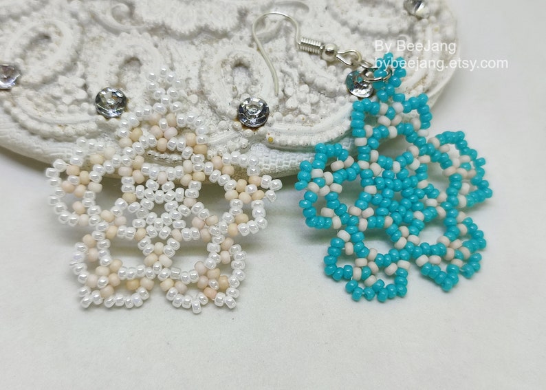 A pair of intricate, handcrafted jewelry pieces, likely earrings, featuring a snowflake or floral design made of seed beads; one is predominantly white/cream colored and the other is turquoise blue and white, displayed on a white background with lace accents.