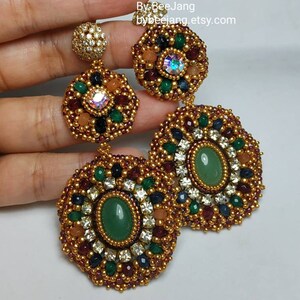 A large pair of intricate, handcrafted drop earrings featuring oval green center stones and bead embroidery in gold, red, dark blue, white, green, and peach colors.