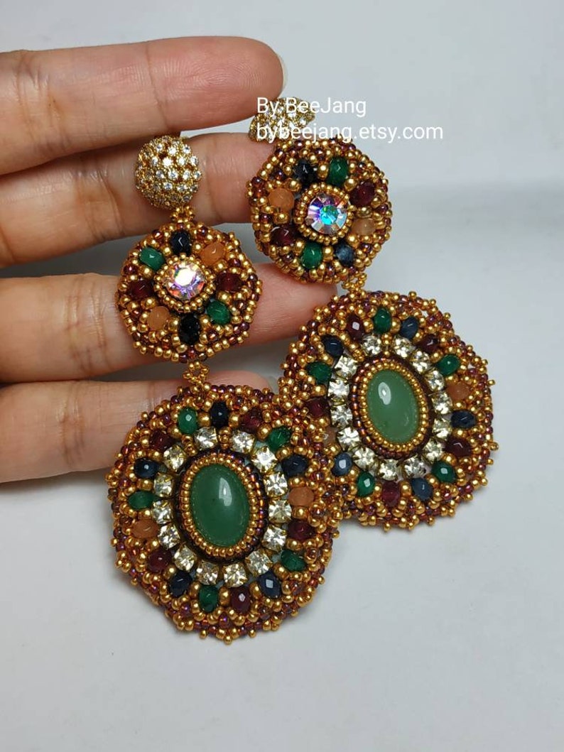 A large pair of intricate, handcrafted drop earrings featuring oval green center stones and bead embroidery in gold, red, dark blue, white, green, and peach colors.