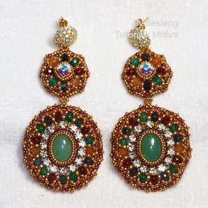 A pair of large, intricate, handcrafted drop earrings featuring an oval green aventurine center stone and extensive bead embroidery using gold, red, dark blue, green, white, and peach-colored seed beads and crystals, displayed on a white surface.