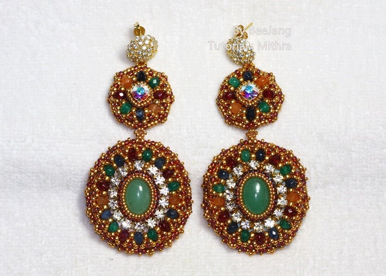 A pair of large, intricate, handcrafted drop earrings featuring an oval green aventurine center stone and extensive bead embroidery using gold, red, dark blue, green, white, and peach-colored seed beads and crystals, displayed on a white surface.