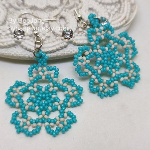 A pair of intricate, handcrafted drop earrings featuring turquoise blue and white seed beads in a snowflake or floral mandala pattern, displayed on a white lace background with small rhinestone details.