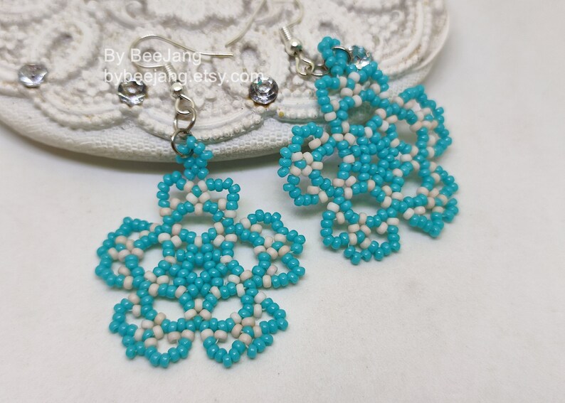 A pair of intricate, handcrafted drop earrings featuring turquoise blue and white seed beads in a snowflake or floral mandala pattern, displayed on a white lace background with small rhinestone details.