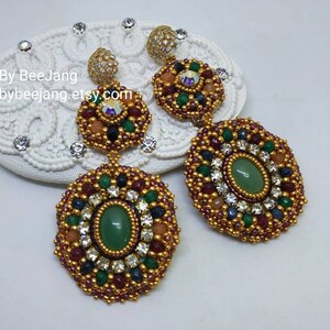 A pair of large, intricate, handcrafted drop earrings featuring an oval green aventurine center stone and extensive bead embroidery using gold, red, dark blue, green, white, and peach-colored seed beads and crystals, displayed on a white decorative surface.