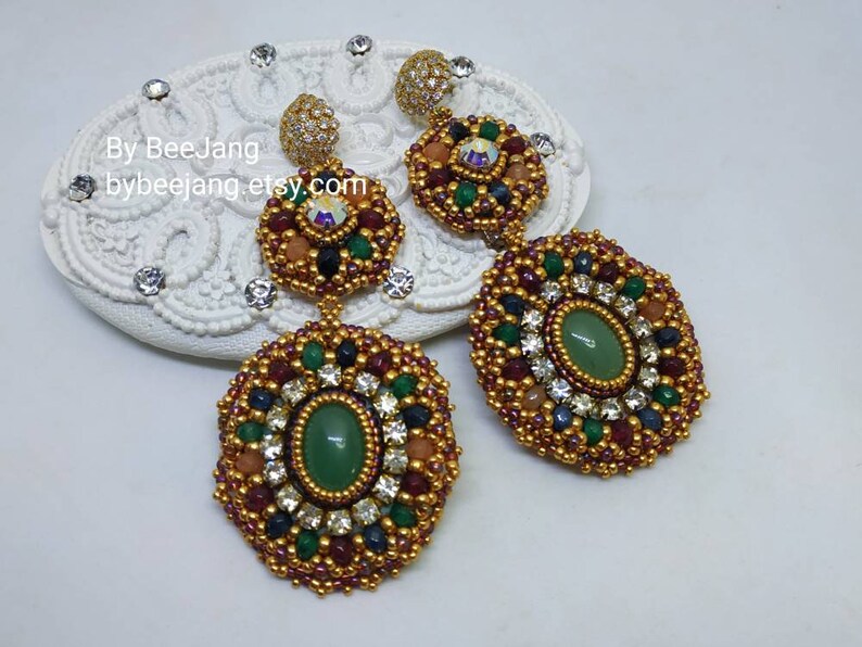 A pair of large, intricate, handcrafted drop earrings featuring an oval green aventurine center stone and extensive bead embroidery using gold, red, dark blue, green, white, and peach-colored seed beads and crystals, displayed on a white decorative surface.