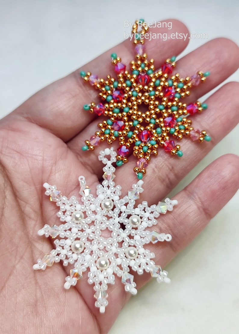 Two handcrafted, intricate beaded snowflake pendants or ornaments being held in a person's hand. The top snowflake features gold, pink/magenta, and turquoise/teal beads, while the bottom snowflake features white, clear, and cream faux pearls/beads.