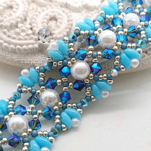 May include: A detailed beaded bracelet in shades of blue and silver. The bracelet features light blue petal-shaped beads, dark blue crystals, and pearl-like beads. The intricate design is set against a white background, showcasing the craftsmanship.
