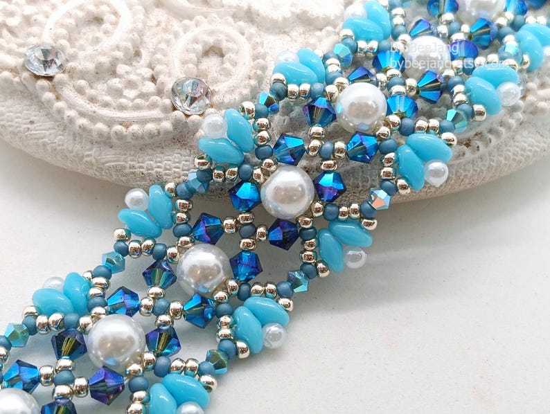 May include: A detailed beaded bracelet in shades of blue and silver. The bracelet features light blue petal-shaped beads, dark blue crystals, and pearl-like beads. The intricate design is set against a white background, showcasing the craftsmanship.