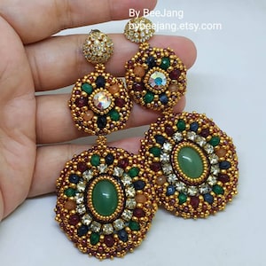 A large pair of intricate, handcrafted drop earrings featuring oval green center stones and bead embroidery in gold, red, dark blue, white, green, and peach colors.