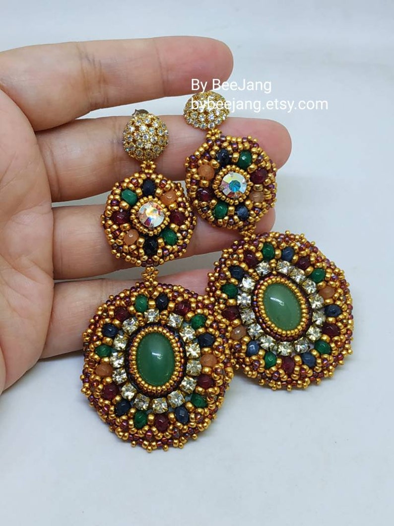 A large pair of intricate, handcrafted drop earrings featuring oval green center stones and bead embroidery in gold, red, dark blue, white, green, and peach colors.