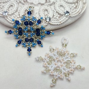Two handcrafted, intricate beaded snowflake pendants or ornaments, one in a blue, silver, and gold color scheme, and the other in a white, cream, and clear palette, displayed on a decorative white lacy surface.