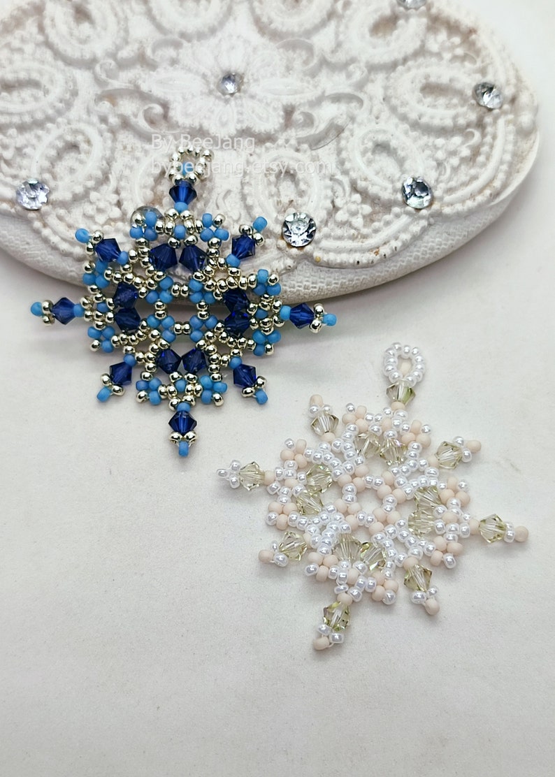 Two handcrafted, intricate beaded snowflake pendants or ornaments, one in a blue, silver, and gold color scheme, and the other in a white, cream, and clear palette, displayed on a decorative white lacy surface.