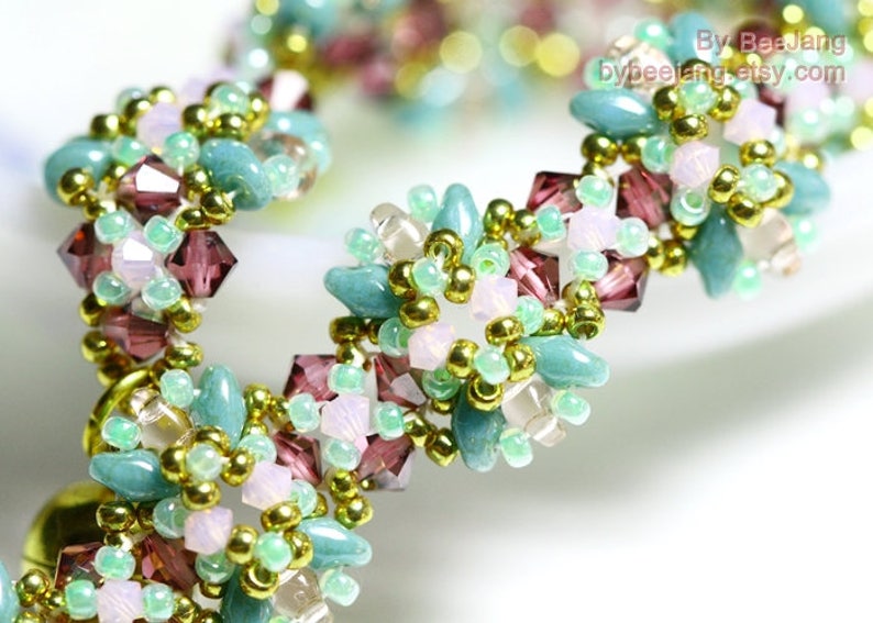 May include: A beaded bracelet featuring a floral design. The bracelet is made with teal, light pink, and purple beads, accented with gold-colored beads. The beads are arranged in a repeating pattern.