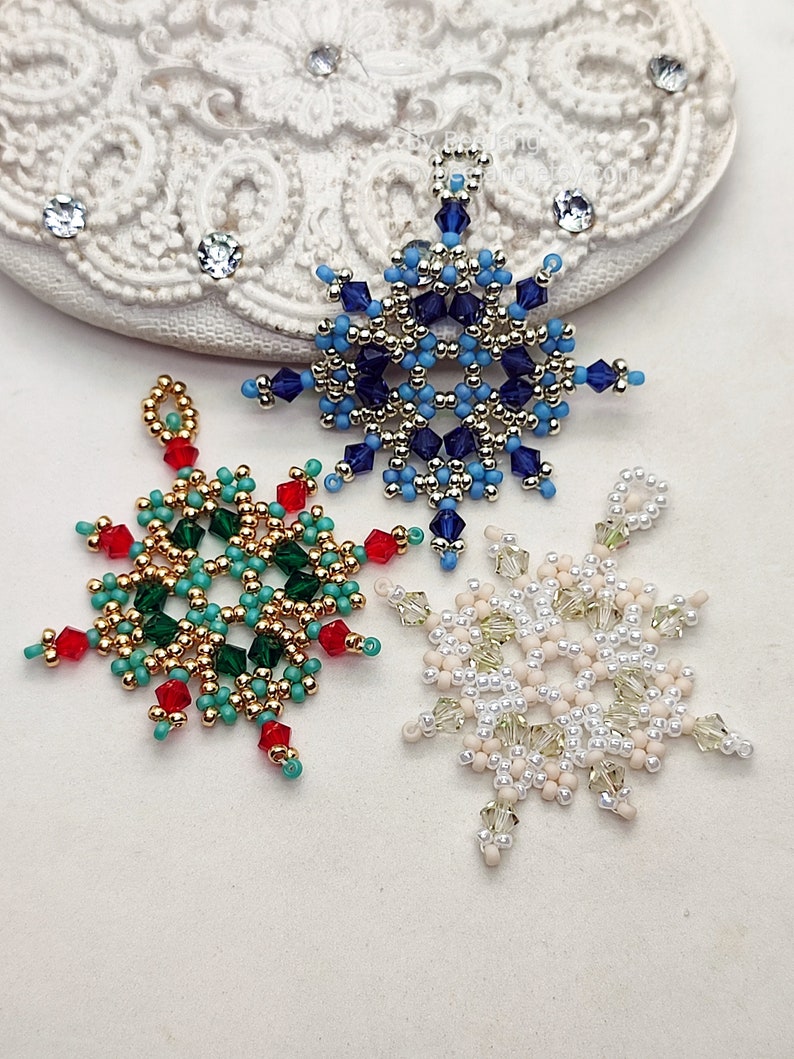 Three handcrafted, intricate beaded snowflake pendants or ornaments, featuring designs in blue/gold/silver, green/gold/turquoise/red, and white/cream/clear color palettes, displayed on an ornate white surface.