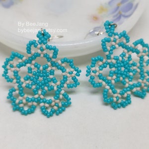 A pair of intricate, handcrafted drop earrings featuring turquoise blue and white seed beads in a snowflake or floral mandala pattern, displayed on a white lace background with small rhinestone details.