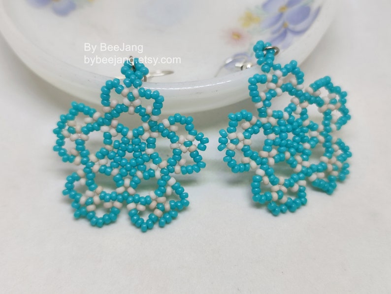 A pair of intricate, handcrafted drop earrings featuring turquoise blue and white seed beads in a snowflake or floral mandala pattern, displayed on a white lace background with small rhinestone details.