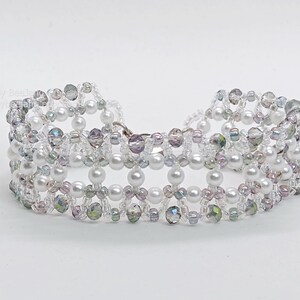 A wide, handcrafted beaded cuff bracelet featuring an intricate, open-lattice
Right Angle Weave design. The piece is constructed with lustrous white pearls, faceted iridescent smoky-grey bicone crystals, and delicate clear and silver-lined seed beads. The beads are woven into a structured, architectural pattern with a shimmering, textured finish, set against a clean white background.