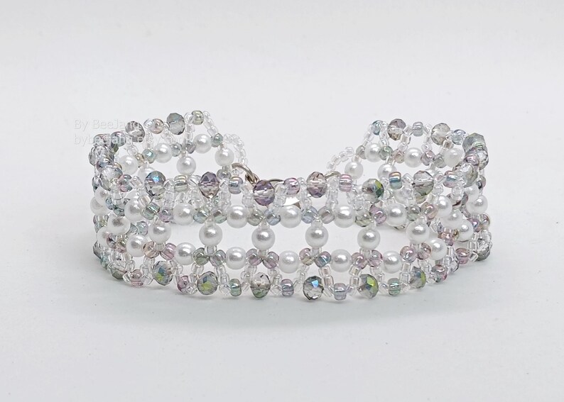 A wide, handcrafted beaded cuff bracelet featuring an intricate, open-lattice
Right Angle Weave design. The piece is constructed with lustrous white pearls, faceted iridescent smoky-grey bicone crystals, and delicate clear and silver-lined seed beads. The beads are woven into a structured, architectural pattern with a shimmering, textured finish, set against a clean white background.