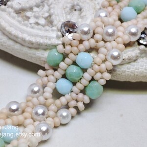May include: A multi-strand beaded bracelet with white, light blue, and green beads. The bracelet features pearl-like beads and is woven in a complex pattern.