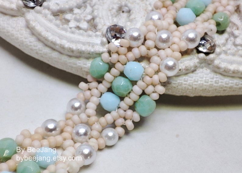 May include: A multi-strand beaded bracelet with white, light blue, and green beads. The bracelet features pearl-like beads and is woven in a complex pattern.