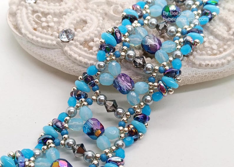 Puede incluir: Una pulsera de cuentas con una mezcla de cuentas azules, moradas y plateadas. La pulsera tiene un dise&ntilde;o en capas con varias formas y tama&ntilde;os de cuentas. El texto "by BeeJann" y "bybeejann.etsy.com" es visible.