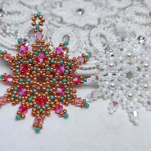 Two handcrafted, intricate beaded snowflake pendants or ornaments displayed on an ornate white lacy surface. The left snowflake features gold, red, turquoise, and pink beads, while the right snowflake features white, clear, and cream faux pearls/beads.