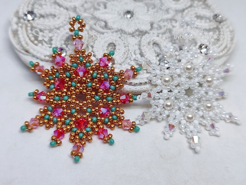 Two handcrafted, intricate beaded snowflake pendants or ornaments displayed on an ornate white lacy surface. The left snowflake features gold, red, turquoise, and pink beads, while the right snowflake features white, clear, and cream faux pearls/beads.