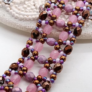 A close-up photograph displays a wide, handcrafted beaded bracelet with a detailed open-lattice pattern. The design uses a combination of faceted light pink bicones, textured lavender round beads, and metallic bronze pearls. These larger beads are interconnected by intricate loops of iridescent purple, white, and bronze seed beads, creating a complex, textured floral effect. The piece is set against a neutral, off-white background that highlights its rich colors.