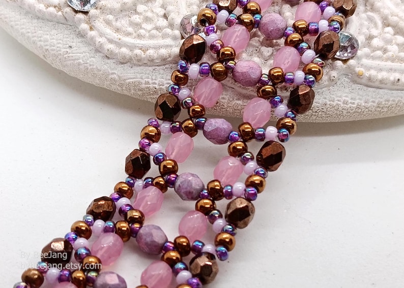 A close-up photograph displays a wide, handcrafted beaded bracelet with a detailed open-lattice pattern. The design uses a combination of faceted light pink bicones, textured lavender round beads, and metallic bronze pearls. These larger beads are interconnected by intricate loops of iridescent purple, white, and bronze seed beads, creating a complex, textured floral effect. The piece is set against a neutral, off-white background that highlights its rich colors.