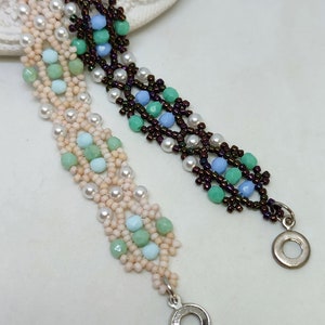 May include: Two beaded bracelets with a silver clasp. One bracelet is made with brown, white, light blue, and green beads. The other bracelet is made with white, light pink, light blue, and green beads. The bracelets are on a white surface.