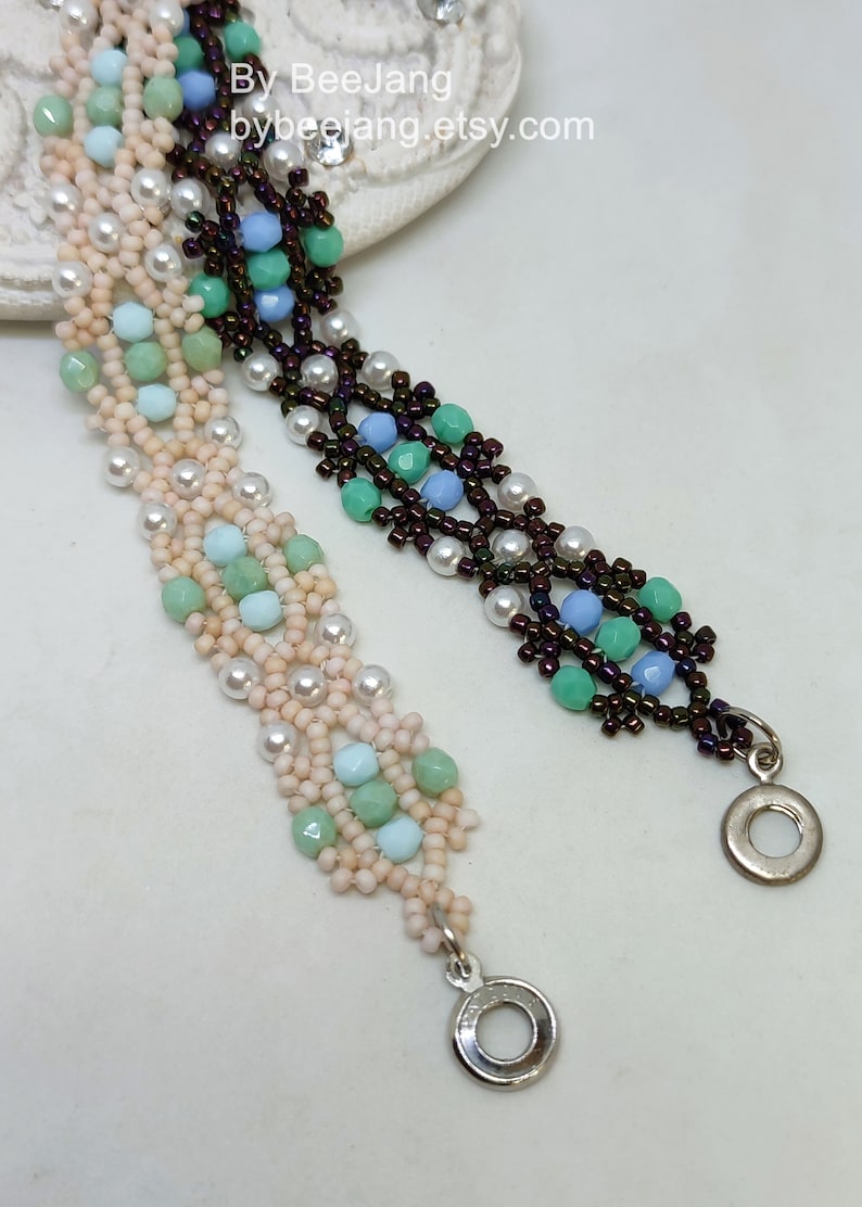 May include: Two beaded bracelets with a silver clasp. One bracelet is made with brown, white, light blue, and green beads. The other bracelet is made with white, light pink, light blue, and green beads. The bracelets are on a white surface.