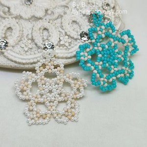 A pair of intricate, handcrafted jewelry pieces, likely earrings, featuring a snowflake or floral design made of seed beads; one is predominantly white/cream colored and the other is turquoise blue and white, displayed on a white background with lace accents.