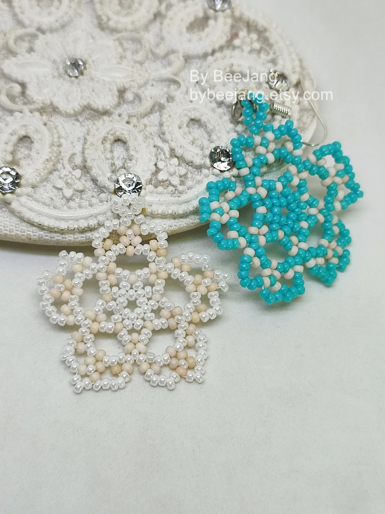 A pair of intricate, handcrafted jewelry pieces, likely earrings, featuring a snowflake or floral design made of seed beads; one is predominantly white/cream colored and the other is turquoise blue and white, displayed on a white background with lace accents.