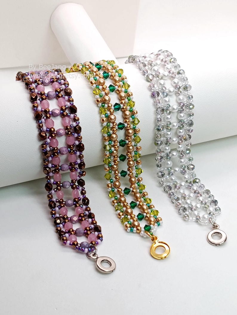 Three wide, handcrafted cuff bracelets are displayed on a white stand, showcasing an intricate Right Angle Weave lattice design. Each piece is meticulously woven with faceted bicone crystals, round pearls, and delicate seed beads, secured with a metallic toggle clasp.

    Left: Purple and lavender crystals with dark metallic bronze accents.
    Center: Emerald and olive green crystals with gold-toned pearls.
    Right: Clear crystals with lustrous white pearls.