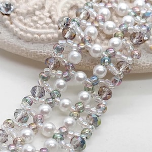 A close-up photo shows a wide, handcrafted beaded bracelet with a delicate, open lattice design. It is meticulously woven using white pearls, faceted smoky grey bicone crystals, and shimmering iridescent seed beads. The piece uses a Right Angle Weave technique to create an elegant, textured floral-like pattern.