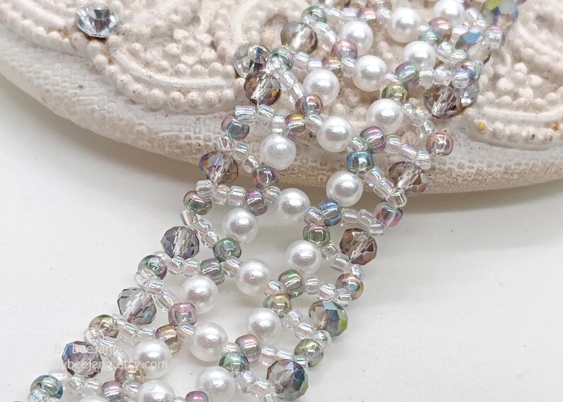 A close-up photo shows a wide, handcrafted beaded bracelet with a delicate, open lattice design. It is meticulously woven using white pearls, faceted smoky grey bicone crystals, and shimmering iridescent seed beads. The piece uses a Right Angle Weave technique to create an elegant, textured floral-like pattern.