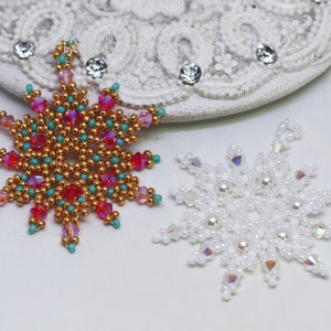 Two handcrafted, intricate beaded snowflake pendants or ornaments displayed on an ornate white lacy surface. The left snowflake features gold, red, turquoise, and pink beads, while the right snowflake features white, clear, and cream faux pearls/beads.