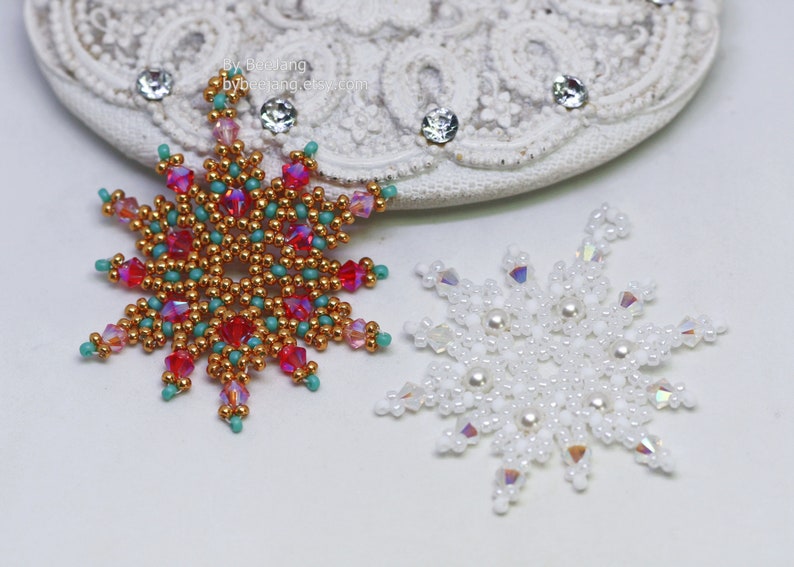 Two handcrafted, intricate beaded snowflake pendants or ornaments displayed on an ornate white lacy surface. The left snowflake features gold, red, turquoise, and pink beads, while the right snowflake features white, clear, and cream faux pearls/beads.