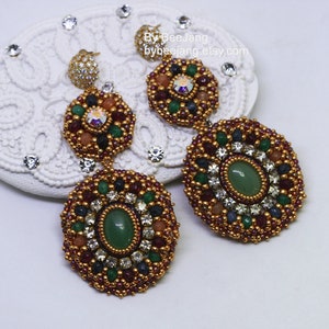 A pair of large, intricate, handcrafted drop earrings featuring an oval green aventurine center stone and extensive bead embroidery using gold, red, dark blue, green, white, and peach-colored seed beads and crystals, displayed on a white decorative surface.