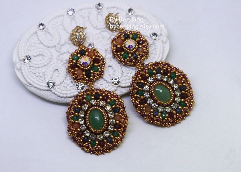 A pair of large, intricate, handcrafted drop earrings featuring an oval green aventurine center stone and extensive bead embroidery using gold, red, dark blue, green, white, and peach-colored seed beads and crystals, displayed on a white decorative surface.