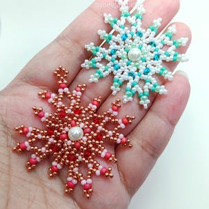 Two handcrafted, intricate beaded snowflake pendants or ornaments being held in a person's hand. The top snowflake features white, clear, and turquoise/blue faux pearls/beads, while the bottom snowflake features copper/gold, red, and pink beads.