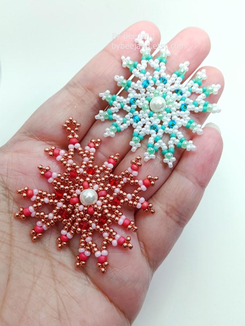 Two handcrafted, intricate beaded snowflake pendants or ornaments being held in a person's hand. The top snowflake features white, clear, and turquoise/blue faux pearls/beads, while the bottom snowflake features copper/gold, red, and pink beads.